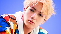 A headshot of Jin a colorful suit and blonde hair