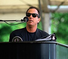 Man plays a piano while singing into microphone. He is wearing dark shades.