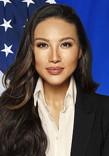 Head shot of Chang in front of the U.S. flag. She is wearing a black suit jacket, white shirt with top button undone, with long brown-highlighted black hair flowing on her right-hand shoulder.