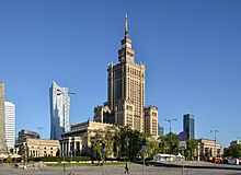 Farbige Eckansicht eines mehrstöckigen, braunen Gebäudes mit einem Turmaufbau und einem Glockenturm. Die Spitze ist mit einem Funkmast versehen. Am großen Turmbau sind bis zur halben Höhe drei Ecktürme angebaut. Auf allen Dächern sind spitze Ornamente angebracht. Links und rechts vom Hauptgebäude sind drei Nebengebäude mit Säuleneingängen, die teilweise von Bäumen verdeckt sind.