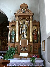 Frontale Farbfotografie eines Altars, das in einer Raumecke steht. Kerzenständer, Topfpflanzen und ein aufgeschlagenes Buch befinden sich vor dem Altar auf einer weißen Tischdecke. Drei Figuren in altertümlicher Kleidung stehen in den Nischen, die von goldenen Säulen verziert sind. Unter und über ihnen sind zwei Porträts mit goldenen Ornamenten.