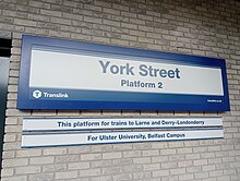 Against a yellow brick wall, a blue-bordered sign with blue text reads "York Street" with "Platform 2" below. There are two smaller signs below that say the destinations of trains to Derry~Londonderry and Larne, as well as to change at the station for the nearby Ulster University campus.