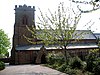 A Gothic Revival sandstone church seen from the side with a crenellated tower on the left and the body of the church with a clerestory to the right; in the foreground on the right is a path with shrubs and a tree that partly obstruct the view of the church