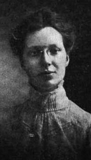 A young white woman with dark hair in an updo, wearing glasses and a high-collared lace-trimmed blouse or dress