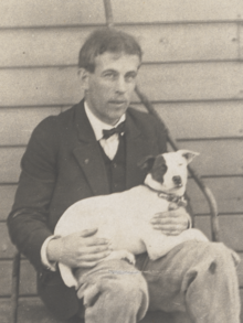 James Arbuthnot, sitting on a sod roller. He's dressed in rough trousers, and coat over a white shirt and vest. His dog is laying across his lap, hind legs on Arbuthnot's right leg. The dog is white save for a dark patch around its right eye.