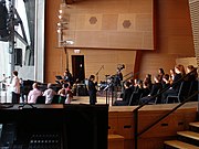 At right a woman in black stands in front of many children dressed in black, sitting in three rows of chairs on a large stage. In the background musicians play a grand piano, drums and guitars. At left a woman stands at a microphone, with several adults seated nearby. The stage opens to the outside at far left.