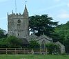 Gray stone building with arched windows. Square tower topped with spirelet, flagpole and weather vane. Foreground has small trees and bushes and a wooden rail fence.