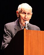 Balding, grey-haired man in his early eighties, standing behind a brown lectern and speaking into a microphone