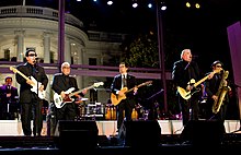 Six male performers playing instruments outdoors, with a background of a white building.