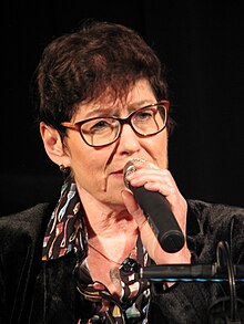 Uschi Brüning ist ein Mikrofon halten au einer Farbfotografie im Hochformat abgebildet. Sie hat kurzes braunes Haar und trägt eine Brille.