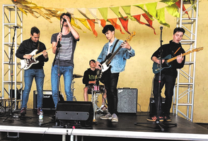 A band consisting of five people plays on a small stage in front of a yellow wall.