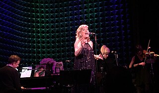 band playing on darkened stage; illuminated at the center is Justin Vivian Bond, a tall femme-presenting sing with blond hair in a lacy black dress