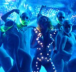 A blond woman dancing in a blue dress, which has small glowing squirrels on it. She is surrounded by dancers in silver, body-hugging dress with a neon green mustard in front