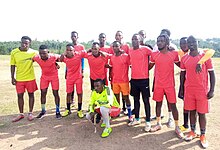 Ein Fußballteam posiert auf einer trockenen Rasenfläche für ein Gruppenfoto. Es sind insgesamt 15 Männer in zwei Reihen stehend, mit dem Torwart vor ihnen hockend. Die erste Reihe hat ihre Arme jeweils über die Schultern der daneben stehenden Teammitglieder gelegt und steht teils leicht nach vorne gebeugt. Im Hintergrund ist ein Waldrand erkennbar.