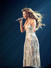 Uçuşan kahverengi saçlı bir kadının sol eliyle sol alt tarafı işaret ettiği, sağ eliyle tuttuğu mikrofona şarkı söylediği bir fotoğraf.