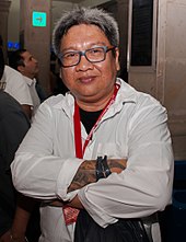 A photograph of a smiling, heavyset gray-haired man wearing glasses with his tattooed arms crossed. Some bystanders are seen in the background.