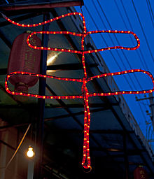 A red LED sign in the shape of two rectangles, one larger than the other, with a line through them, hanging from a building's eave against a darkening sky