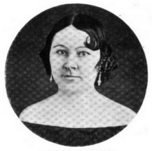 Portrait photograph of a middle-aged woman with her dark hair in an up-do, who is wearing drop earrings and an off-the-shoulder dark blouse.