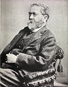 Black and white photo of a seated man with beard and glasses