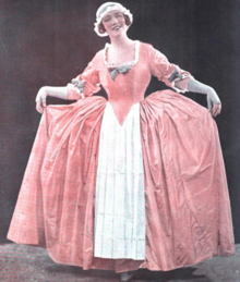 A smiling white woman, standing on stage, wearing an 18th century-style costume, a wide pink dress with a white apron and cap