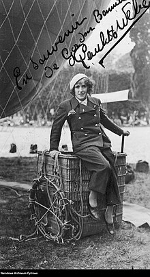 Young woman sitting on the edge of a hot air balloon basket that is set on the ground. She's wearing pants, a double-breasted jacket and a beret. There is a part of a hot air balloon looming behind her.