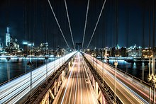 Atmosphärisches Foto der Manhattan Bridge in der Nacht. Blick von einem Brückenpylon in Richtung Manhattan entlang der Fahrbahnen. Die Lichter der auf mehreren Fahrspuren fahrenden Kraftfahrzeuge sind durch die Langzeitbelichtung zu roten und weißen Spuren verwischt. Die Lichter reflektieren von dicken Brückentragseilen, die sich in der oberen Bildhälfte in die Ferne zu erstrecken scheinen. Rechts und links des Brückendecks reflektieren die Lichter der Stadt im dunklen Wasser, im Hintergrund sieht man Hochhäuser und ufernahe Bebauung von Manhattan.