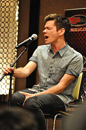 Açık mavi renkte bir gömlek giyen, sarı bir sandalyede oturan ve mikrofona şarkı söyleyen bir adam. Sağ eli ile mikrofonu, sol eli ile bacağını tutuyor.