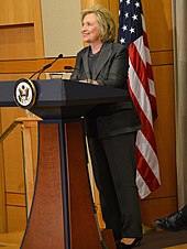 Clinton standing behind lectern wearing a charcoal-colored suit, smiling and looking to her right