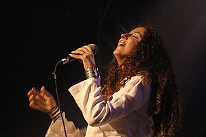 Lynda Thalie wearing a white garment, standing onstage, leaning back and singing into a microphone with eyes closed