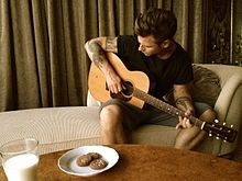 Photograph of Peter Svensson one of "Bad Idea"s composers sitting in a chair playing an acoustic guitar behhind a table containing a glass of milk and a plate of cookies