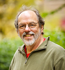 A bearded man in his early sixties grinning while wearing eyeglasses and a fleece jacket