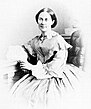 Die Aufnahme der 1860er Jahre zeigt Emma Schenson mit dunklen Haaren, in der Mitte gescheitelt und hinten vermutlich geknotet. Ihr hochgeschlossenes Kleid ist eine Krinoline. Der linke Arm ruht auf einem Gestell mit Säulen.