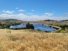 A hillside covered in dry grass overlooks a large shallow lagoon of blue water