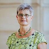 Professor Sigrid Betzelt hat kurze graue Haare mit Mittelscheitel. Sie trägt eine Brille und zu einer mit Umrissen großer Blüten bedruckten sommerlichen Kurzarmbluse in einem hellgrünen Ton eine am Schlüsselbein anliegende Holzperlenkette mit grün-blau-gelben Holzperlen