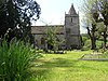 Gray stone building with small square tower and pyramidal roof. Grassy foreground with a cross and gravestones