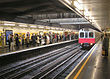 A red and grey train with a black sign that reads "UPMINSTER" in yellow letters is on railway tracks leading from the background to the foreground.