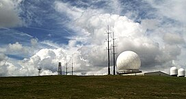 Photo shows a large white golfball-style radar dome, with two smaller radar domes and a number of other antenna; all set against a dramatic cloudy sky.