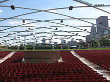 A large number of red seats with a green lawn and park behind, beneath a symmetic curving metal trellis with speakers. Skyscrapers are in the distant background.