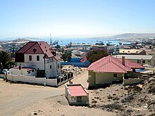 Colour photograph of Lüderitzof taken close to Diamantberg Street. Red tiled houses can be seen in the foreground, and in the background, the Atlantic Ocean can be glimpsed in a bay that is surrounded on sides by rocky land.