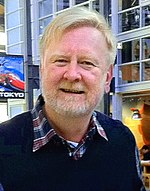 An older man is seen wearing a blue sweatshirt over a collared shirt with a red and blue plaid design.