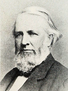 Head and shoulders photograph of a man in an overcoat. He has a full beard with sideburns and has his white hair parted at the his right with it being of enough length to be tucked a few inches behind his left ear. He wears a serious expression and is looking to the left.