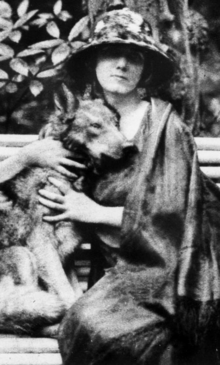 A young white woman wearing a flowery hat with a wide, low brim and a long cloak, seated on a bench outdoors, embracing a dog