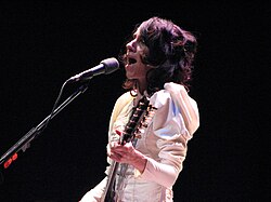 A female musician performing live onstage. She is wearing a white Victorian-style dress and performing a song on an electric guitar. A microphone is in front of the musician.