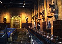 Farbfotografie von einer mittelalterlichen Halle als Kulisse, die mit Absperrseilen durchzogen ist. An den Wänden halten große Figuren Kerzenschalen und im Hintergrund ist ein goldenes Rundbogentor. Auf der ganzen rechten Seite sind eine gedeckte Tafel und drei graue Kleider auf Schneiderbüsten. In der Mitte ist ein Tisch mit einem blauen Tuch und gelben Ringen darauf.
