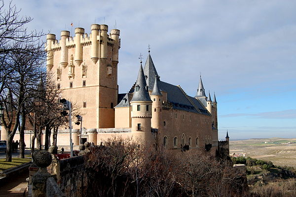 Photo of a Spanish castle high on a rocky peninsular above a plain. It is dominated by a tall rectangular tower rising above a main building with steep slate roof. The walls are pink, and covered with a sculptual pattern. There is a variety of turrets and details.