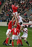 Two rugby players in red shirts and white shorts lift a team-mate into the air to catch a ball, while others dressed in white stretch to intervene.