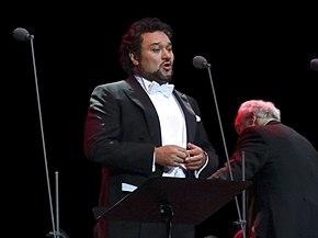 Tenor Ramón Vargas is standing in front of a black backdrop, wearing a tuxedo. Next to him are microphones.