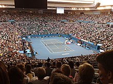 A blue tennis hard court, in the middle of a stadium with crowded bleachers