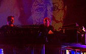 A dark image of two men in the back playing the synthesizers.