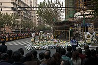 A large crowd stands behind metal fences around the base of a burned-out building, which is surrounded by many flowers and wreaths.